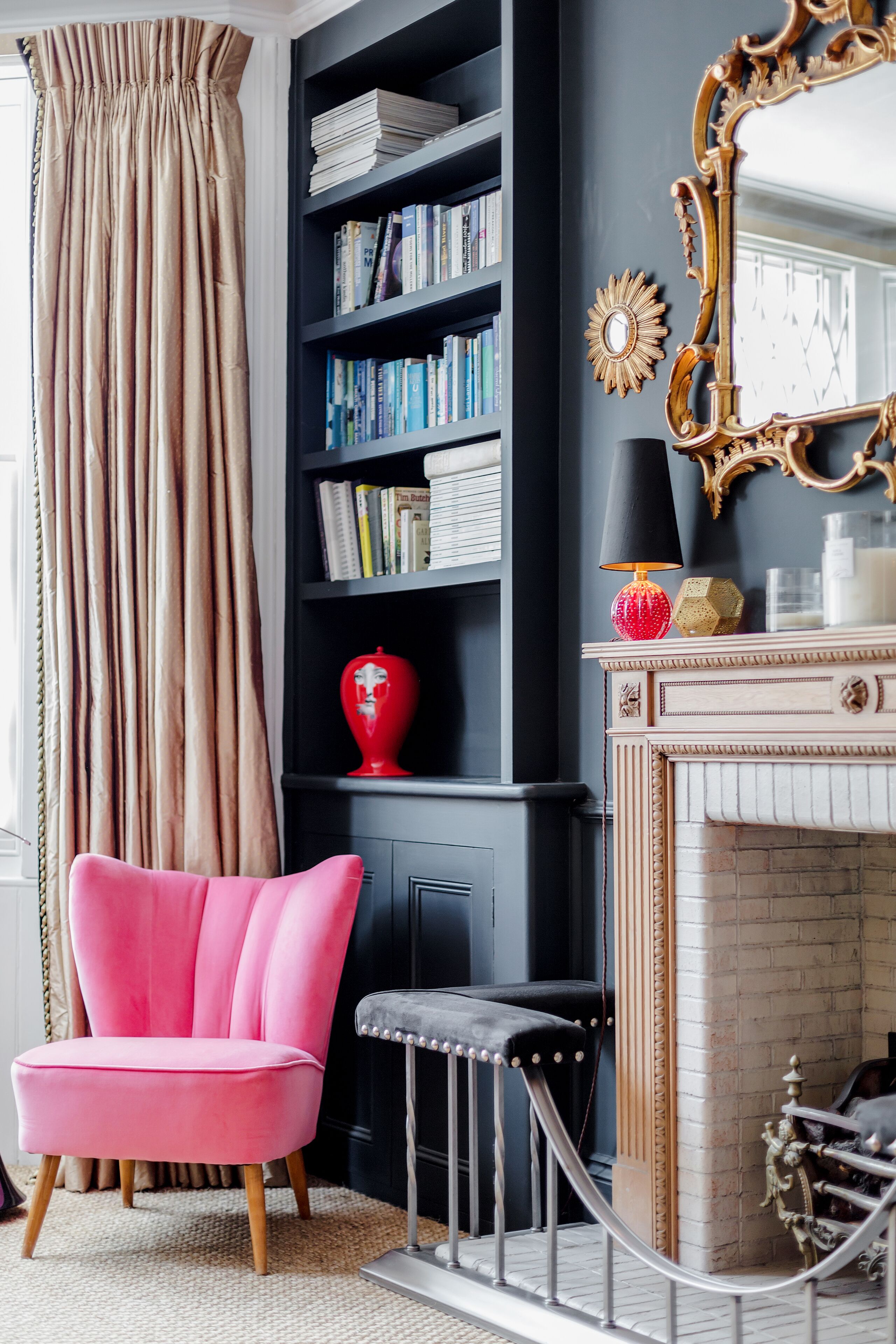 Una habitación con una estantería negra, un jarrón rojo, una chimenea y una silla rosa.