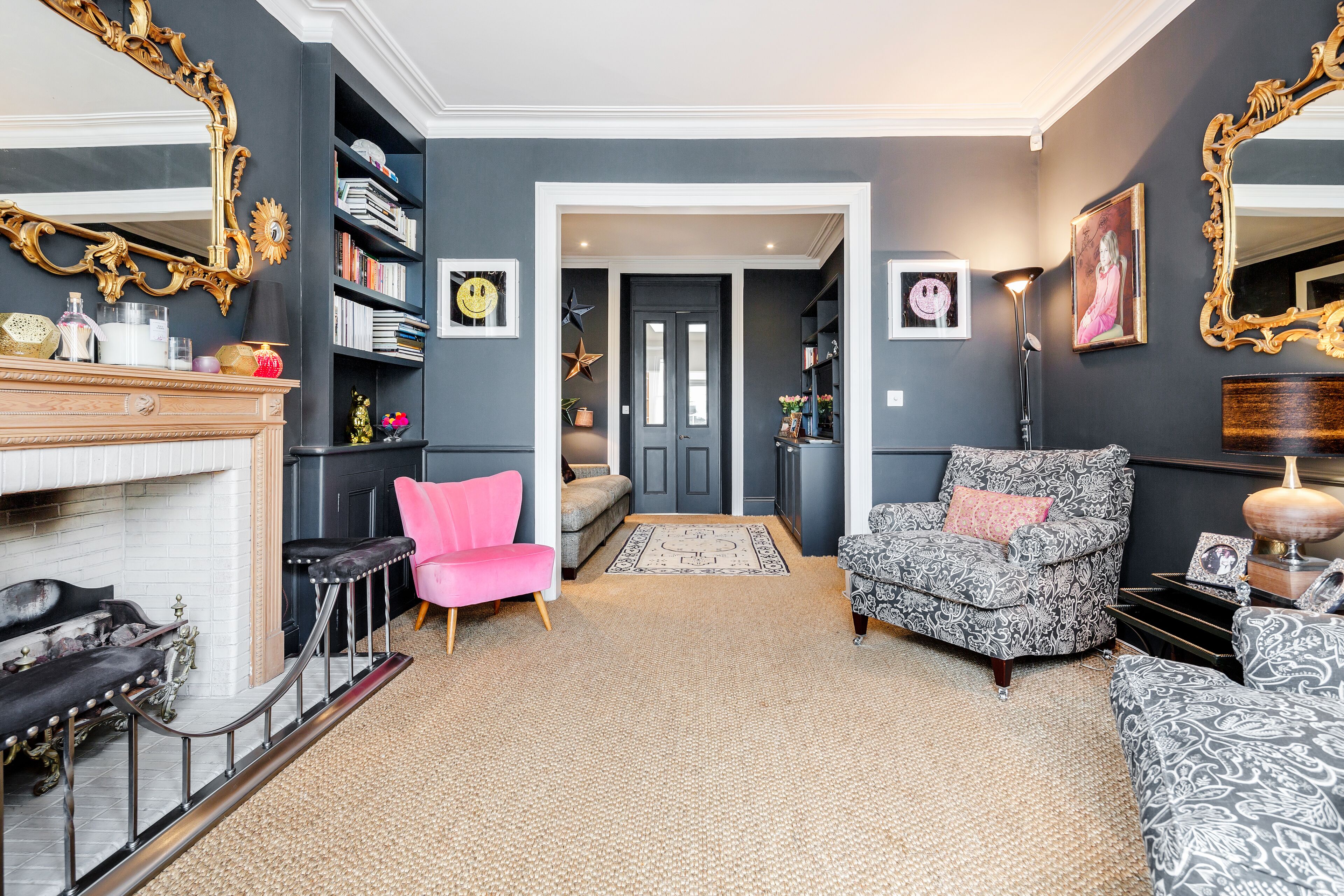 Un salon avec une cheminée, un fauteuil rose, un fauteuil à motifs et une bibliothèque.