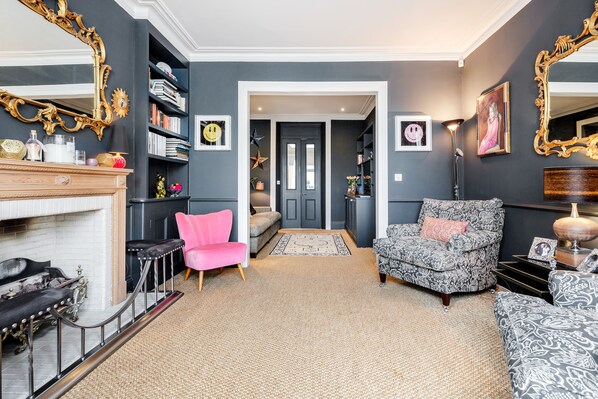 Una sala de estar con chimenea, un sillón rosa, un sillón estampado y una estantería.