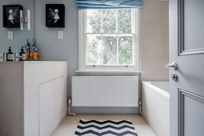 Un baño con un radiador blanco, una ventana con vistas a árboles y un felpudo con diseño en chevron.
