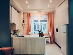 Une cuisine moderne dotée d’un îlot central, de meubles blancs et offrant une vue sur les bâtiments grâce à de grandes fenêtres.
