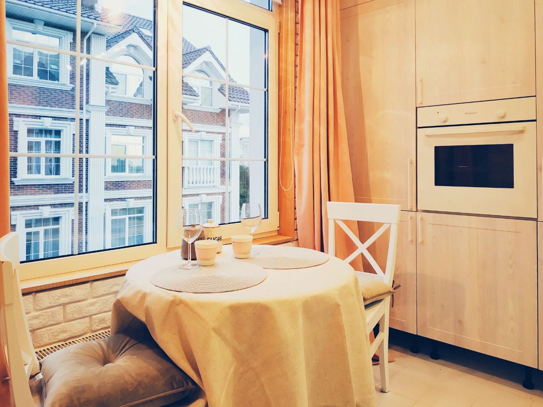 A cozy dining area with a table set for two, a window with a view of a row of houses, and a modern kitchen with a built-in oven.