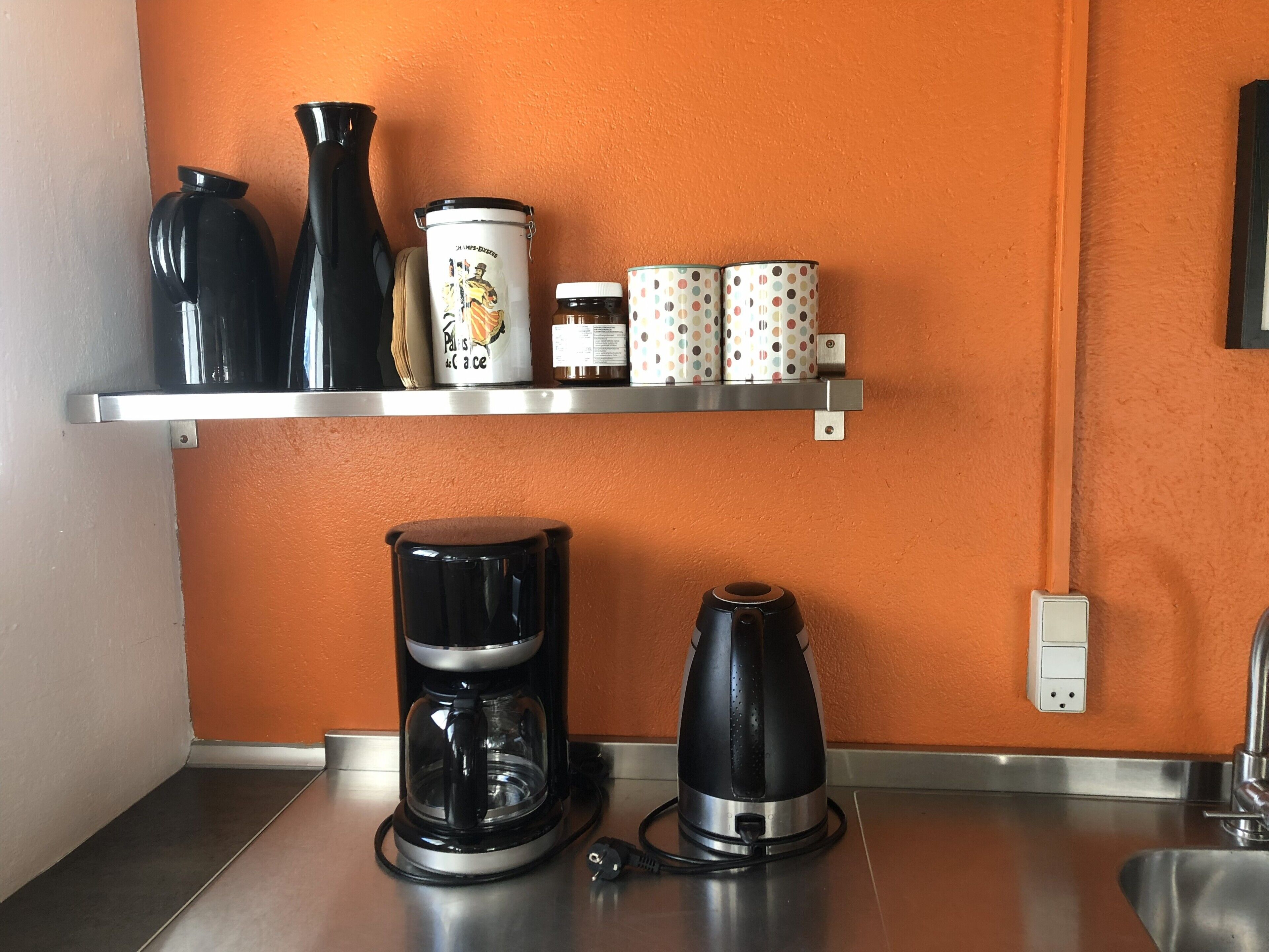 A kitchen with a coffee maker, a kettle, and a shelf with jars and a polka-dotted container.