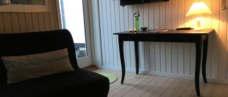 A living room with a black sofa, a small table with a lamp, a TV mounted on the wall, and a window with a view outside.