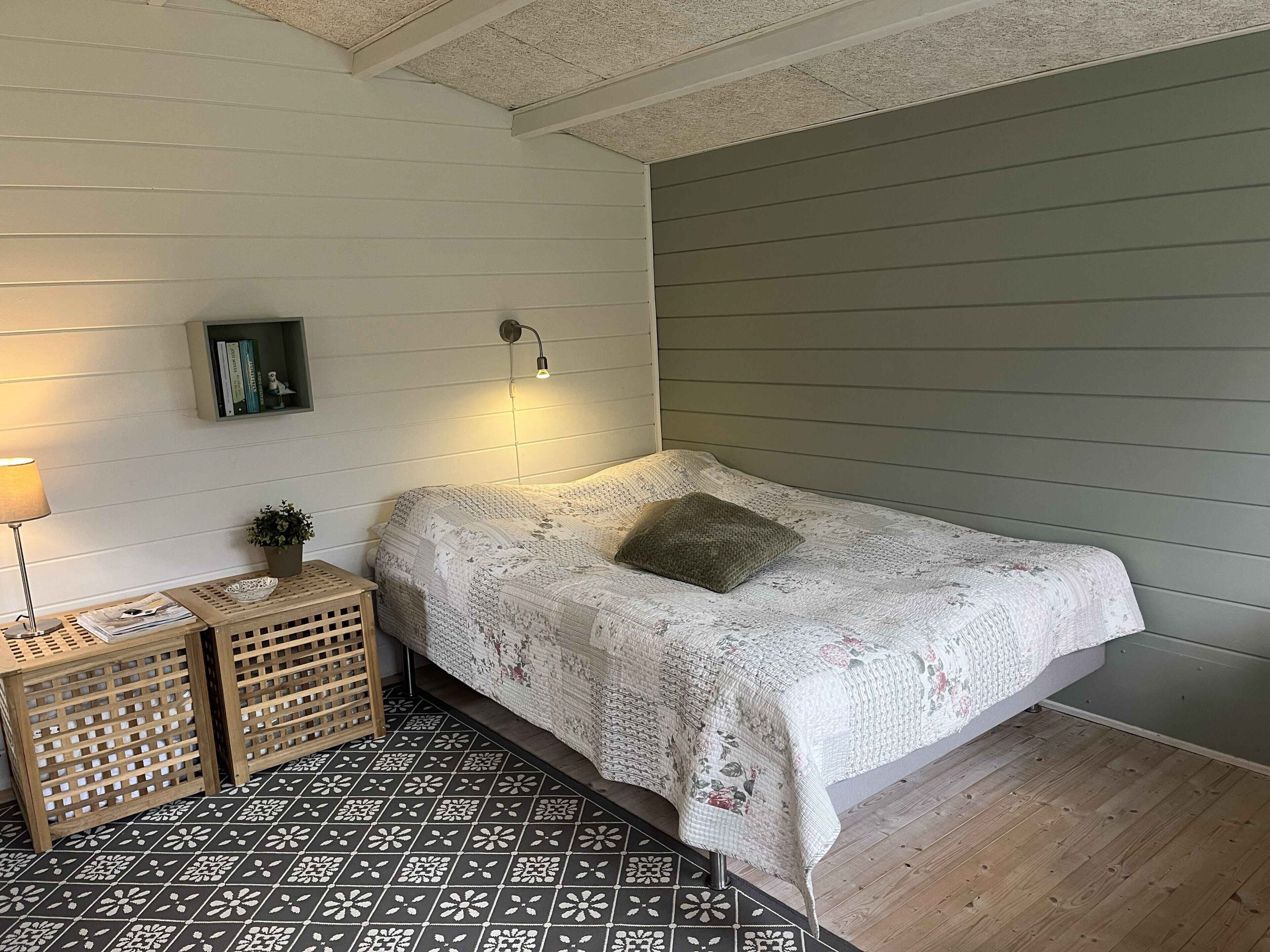 A bedroom with a bed, a nightstand, a lamp, a wall-mounted shelf, and a patterned floor.