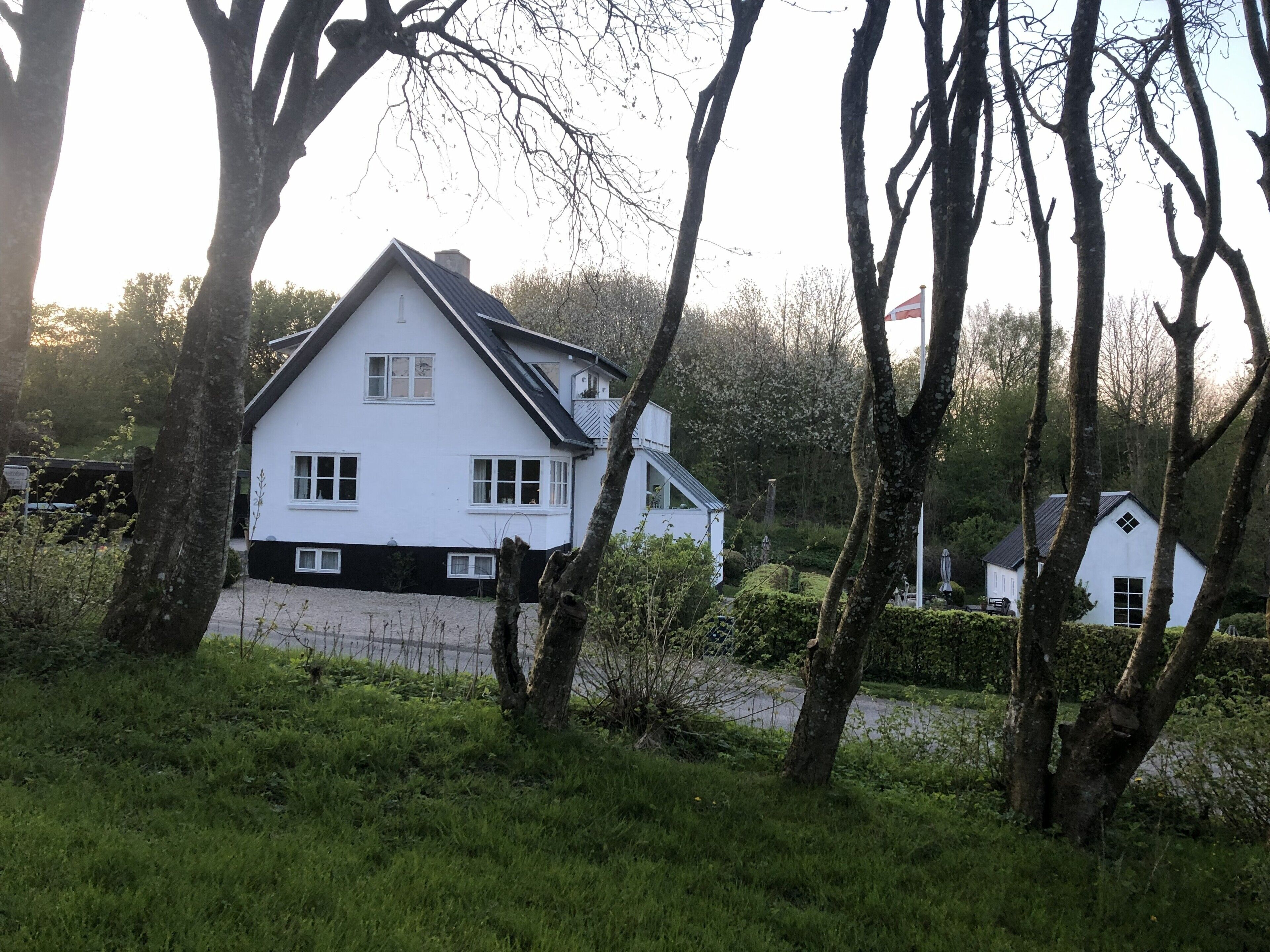 Uma casa branca de dois andares com detalhes em preto na base, cercada por árvores e vegetação.