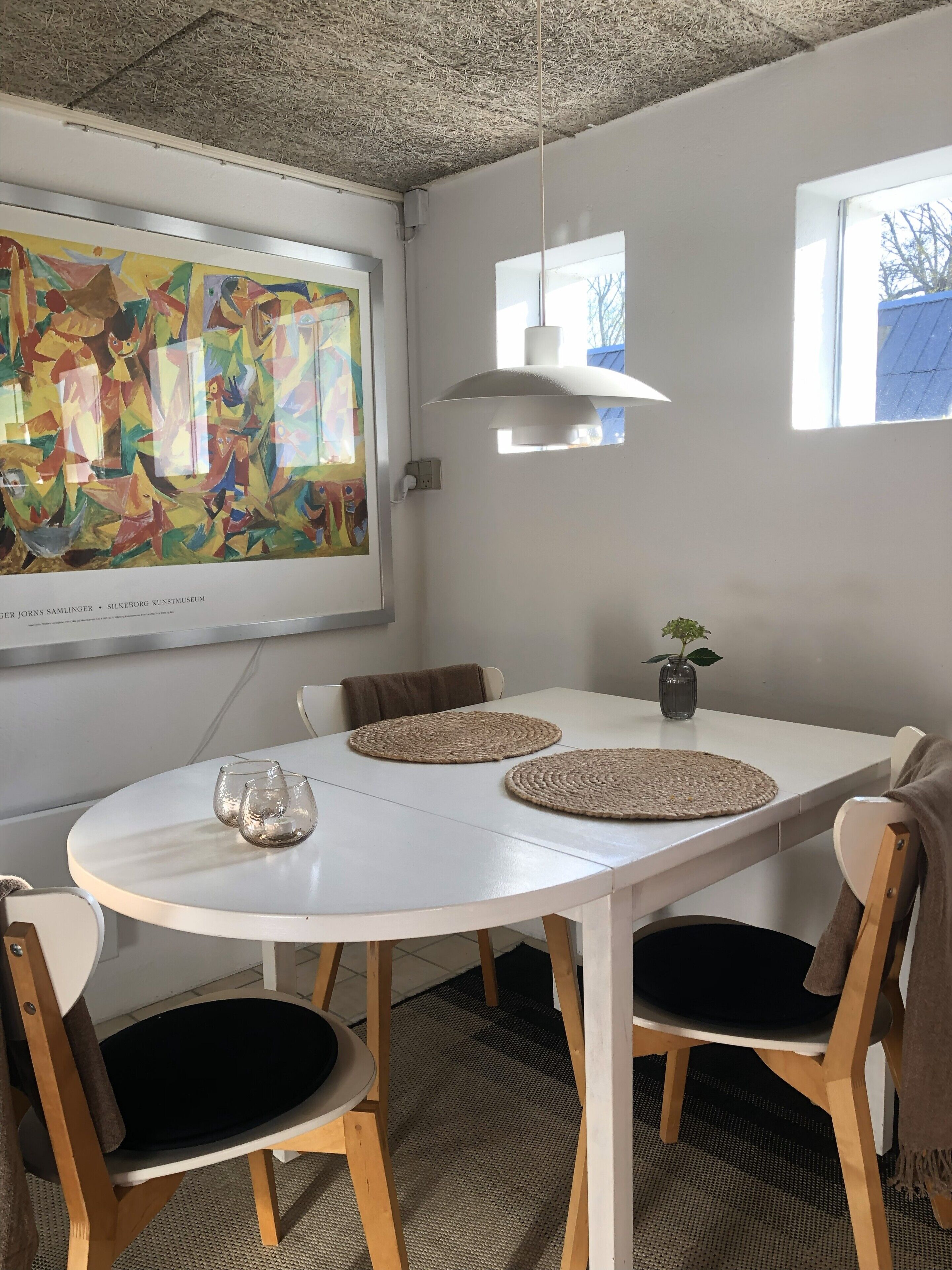 A dining area with a white table, four chairs, a ceiling with a textured pattern, a hanging light fixture, and a colorful abstract painting on the wall.