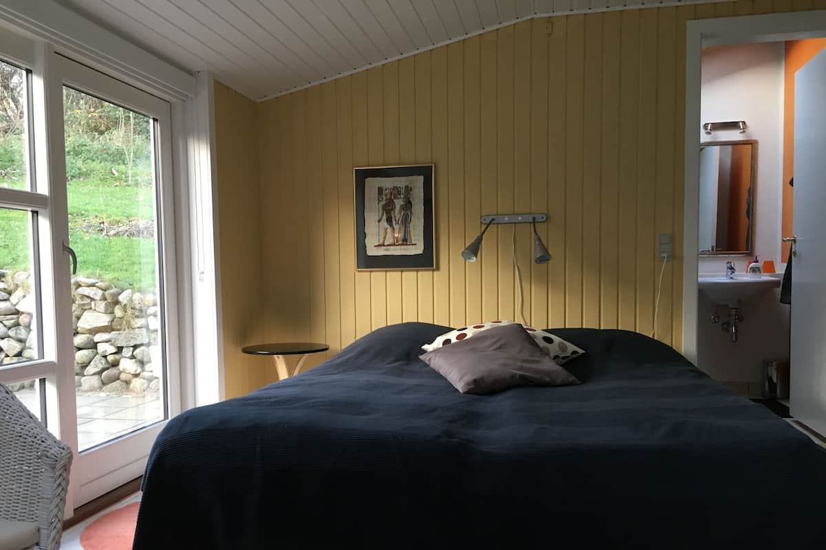 Un dormitorio con una cama, una mesita, un cuadro en la pared y un baño visible a través de una puerta abierta.