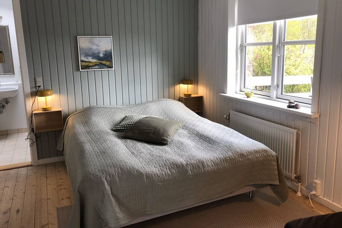 Un dormitorio con cama, lámparas de noche, ventana con vistas y un cuadro en la pared.