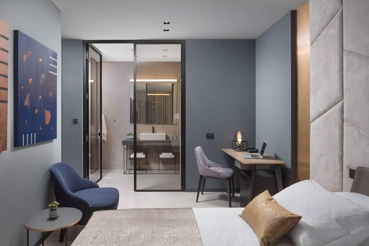 Una habitación de hotel moderna con cama, escritorio, silla, una pequeña mesa y un baño visible a través de una puerta de vidrio.