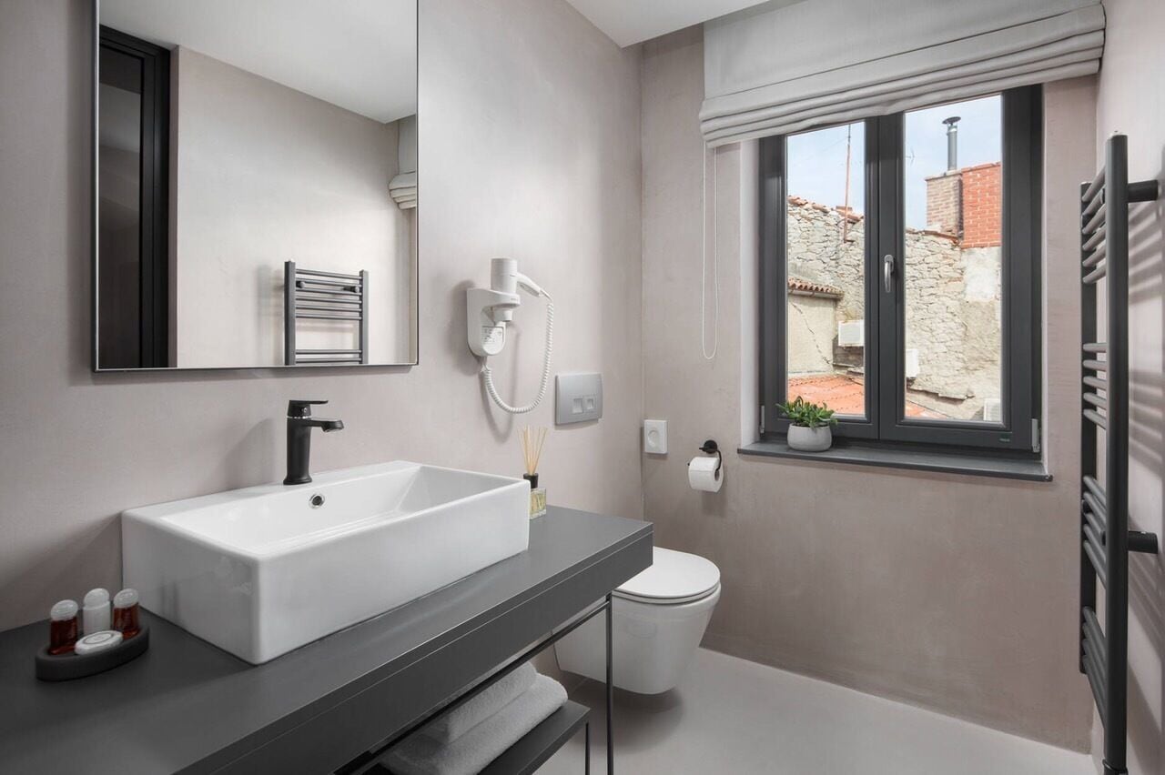 Ein modernes Badezimmer mit einem großen weißen Waschbecken, einer schwarzen Armatur, einem Handtuchhalter, einer Toilette und einem Fenster mit Blick auf Gebäude.