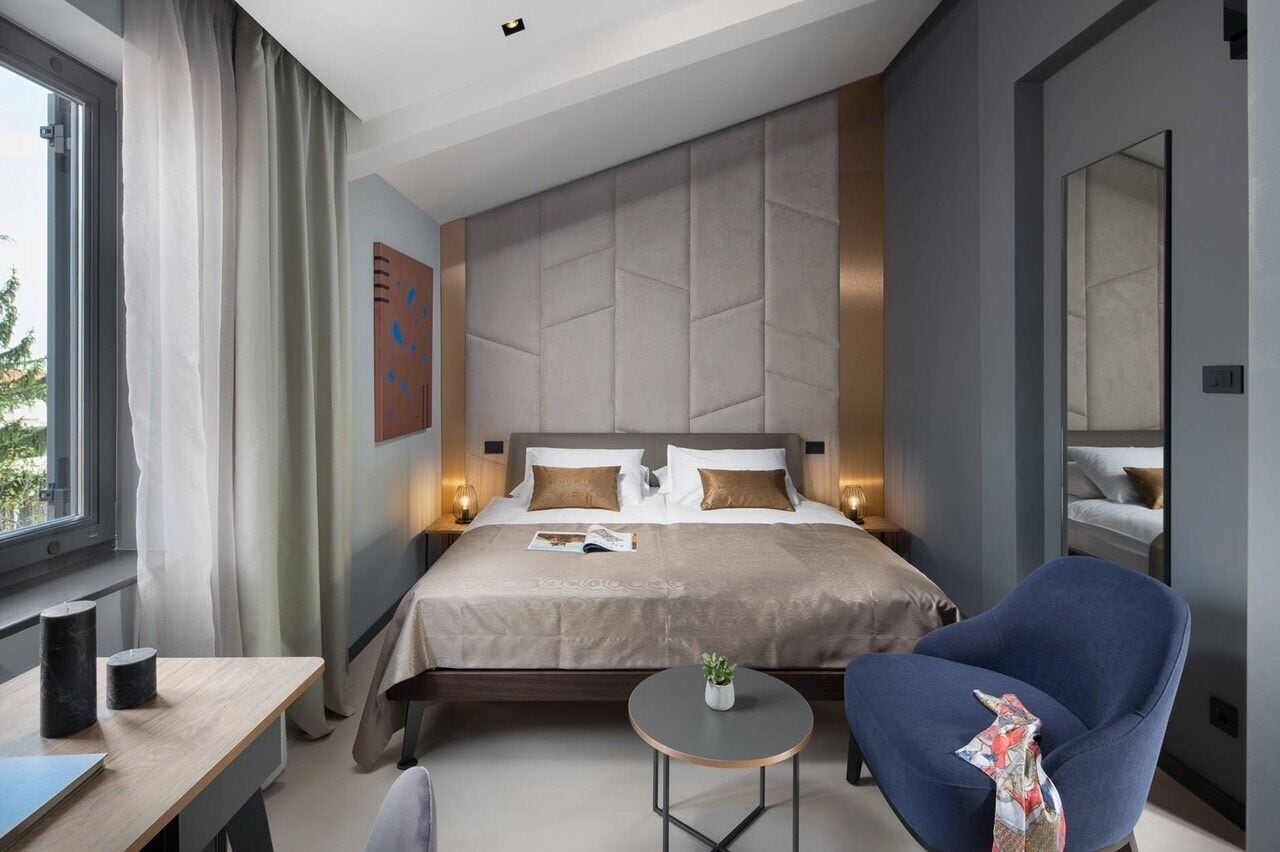 A modern hotel room with a large bed, a bedside table, a chair, a small round table, and a window with curtains.
