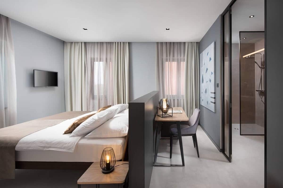 Una habitación de hotel moderna con una cama grande, un escritorio con silla, un televisor en la pared y una zona de ducha.