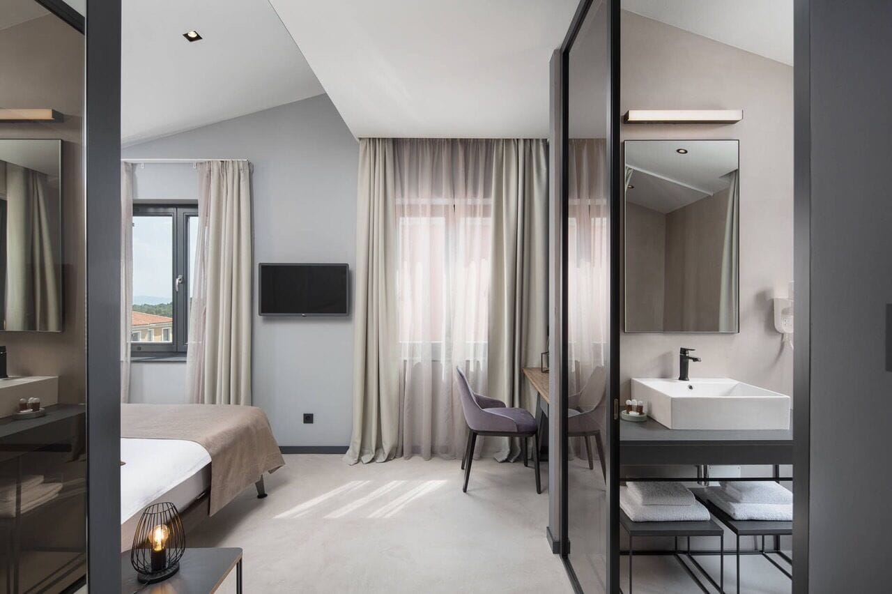 Habitación de hotel moderna con cama, escritorio, silla, espejo grande, televisión y baño con lavamanos y perchero para toallas.