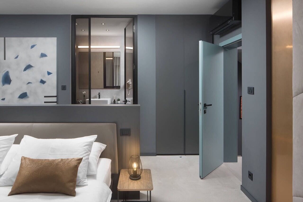 A modern hotel room with a large bed, a bedside table with a lamp, a bathroom with a mirror and sink, and a wardrobe.