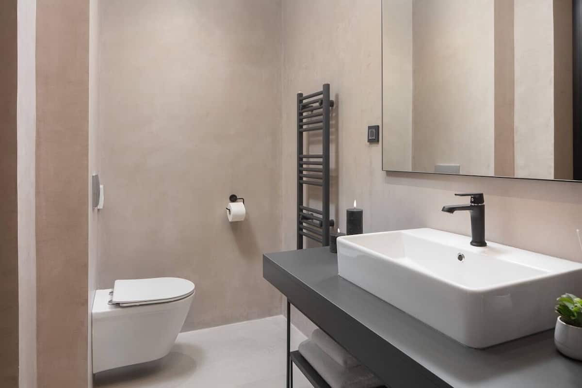 Un baño moderno con inodoro blanco, toallero negro, lavamanos blanco con grifo negro, y un espejo sobre el lavamanos.