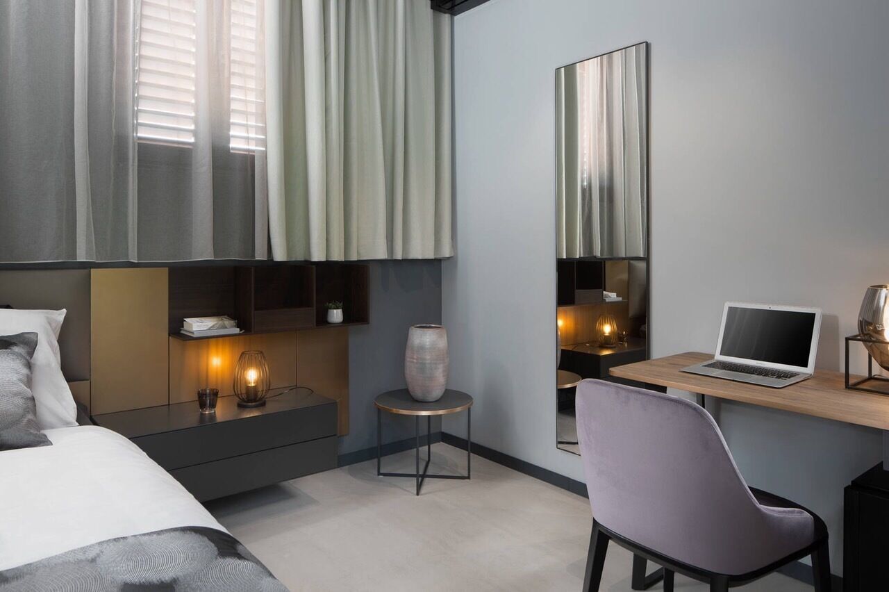 A modern hotel room with a bed, bedside table, lamp, vase, desk with laptop, chair, and large mirror.