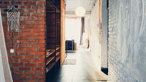 A narrow hallway with a brick wall on one side and a white-painted wall on the other. There are shelves with various items on the left, a hanging light fixture in the center, and a door at the end of the hallway. - MalinaApt  Bohemian Loft (Kyiv)