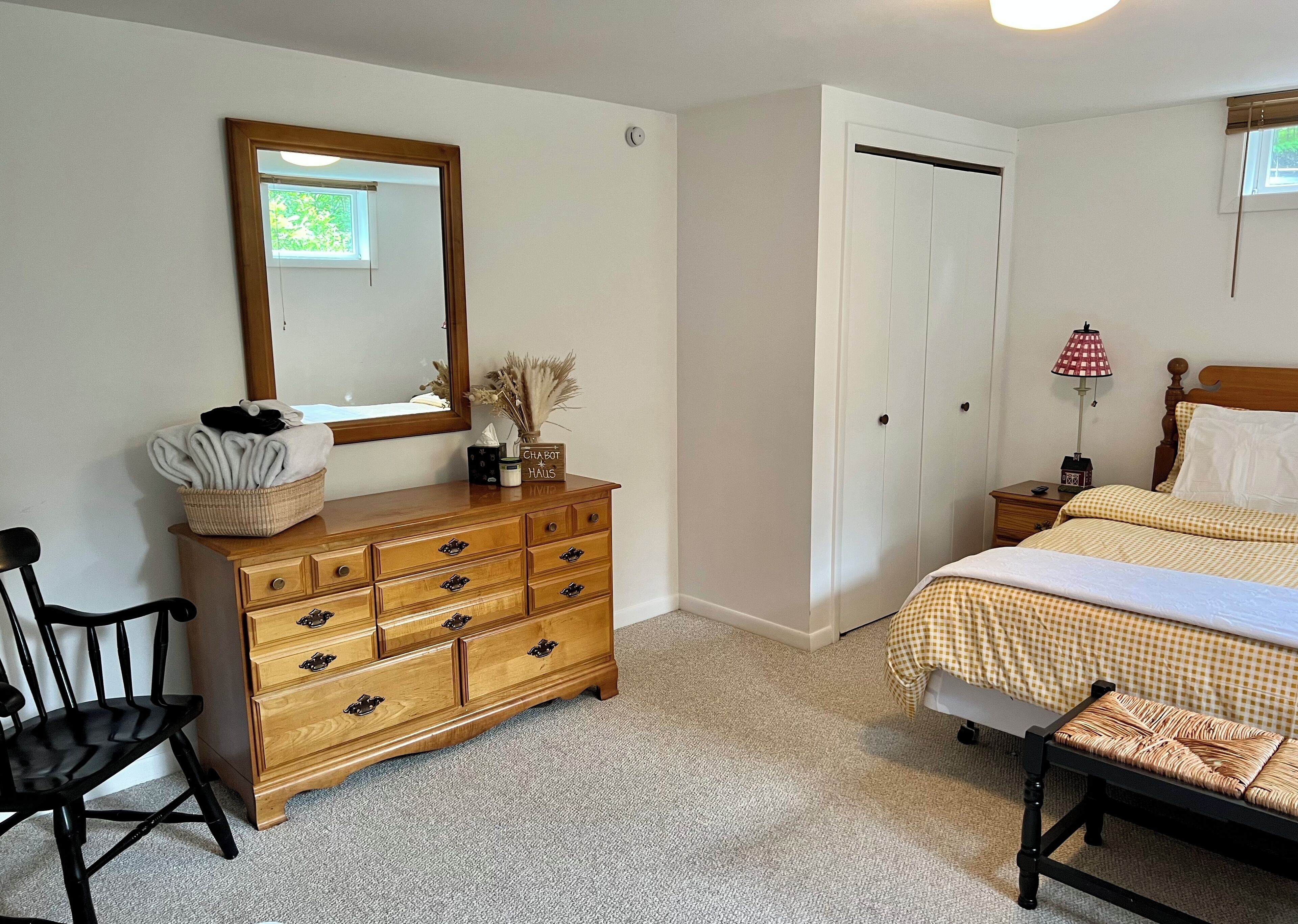 A bedroom with a wooden dresser, a bed with checkered bedding, a nightstand with a lamp, and a chair.