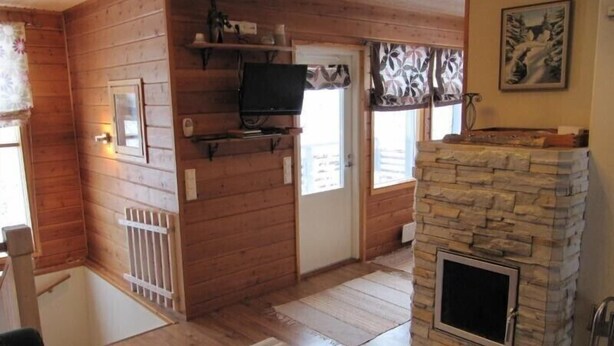 A wooden cabin interior with a stone fireplace, a television mounted on the wall, and a window with a floral curtain. - Rukako JYRKKÄLÄ A (Kuusamo)
