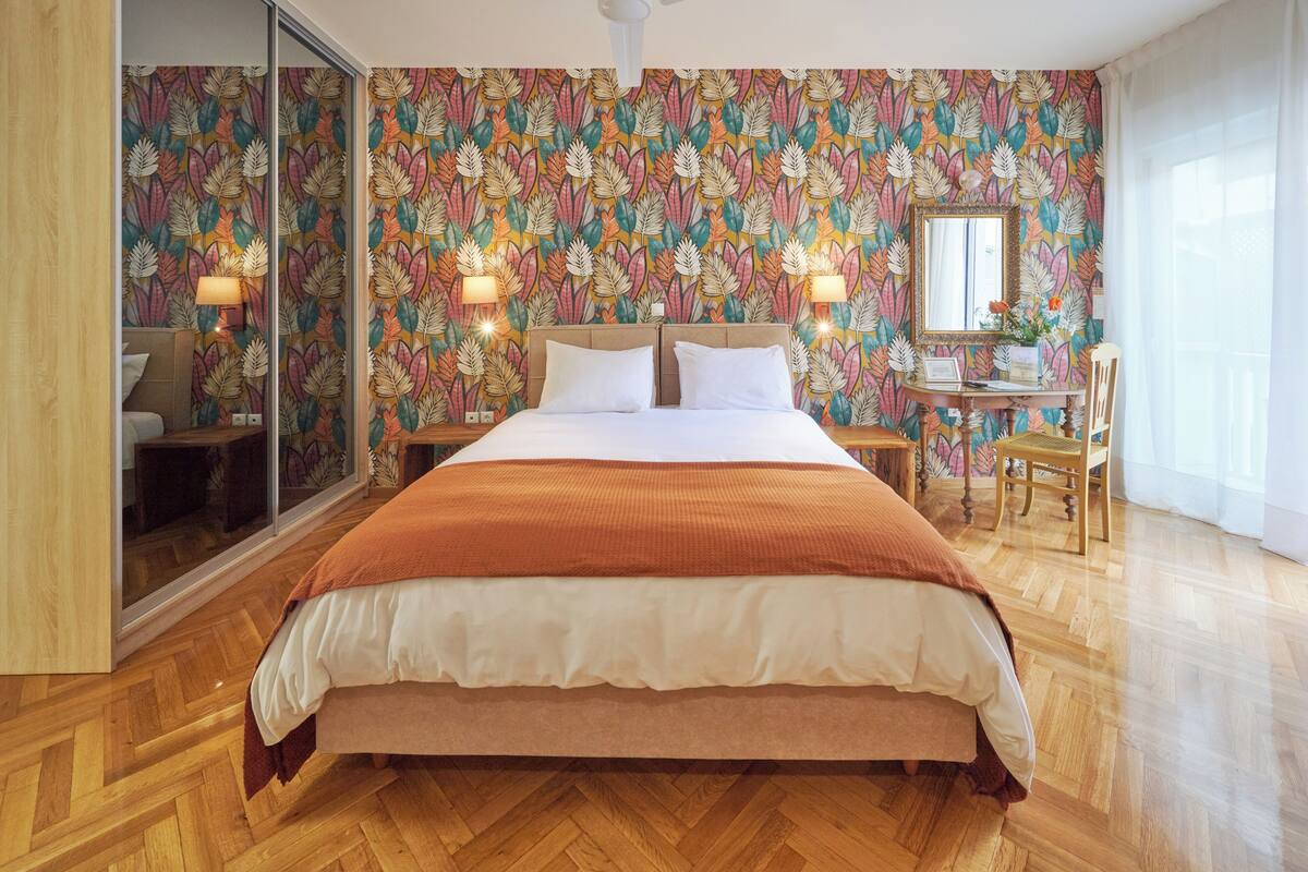 A bedroom with a large bed, wooden flooring, a mirror, and a desk with a chair.