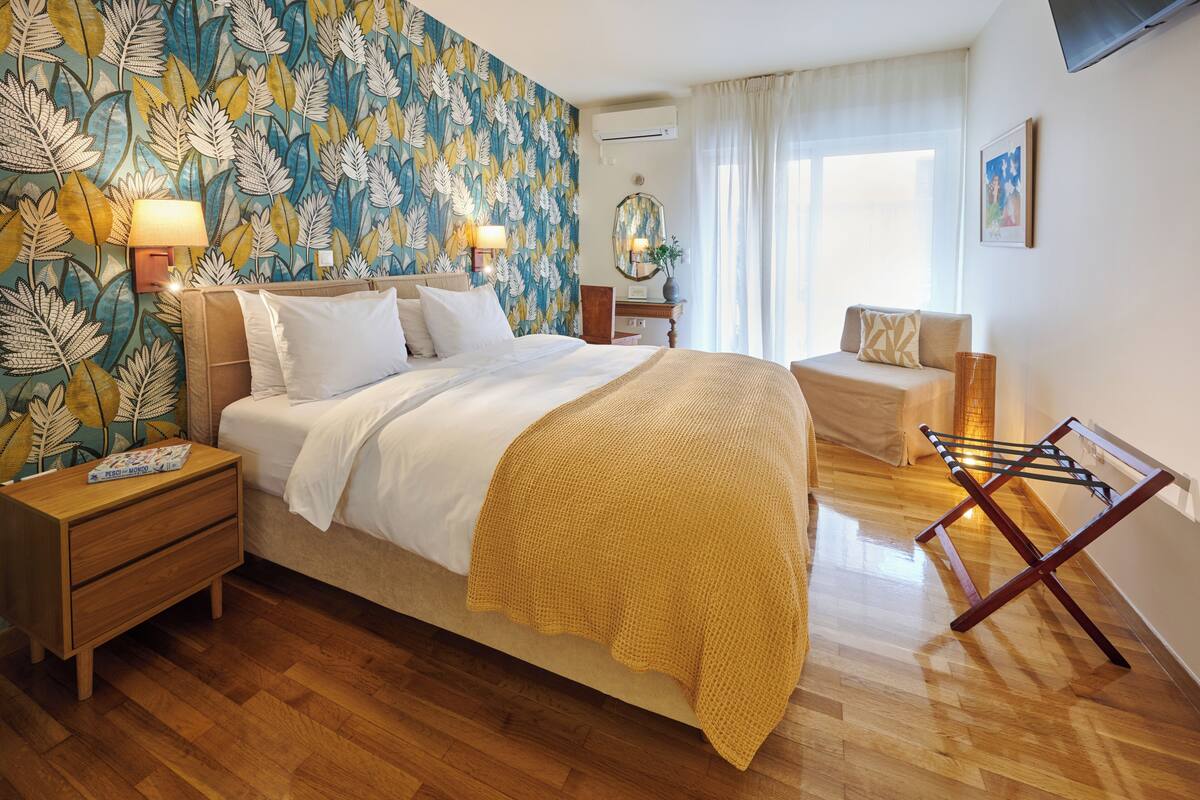 A bedroom with a bed, bedside tables, a nightstand, a mirror, a window with curtains, and a wall-mounted TV.