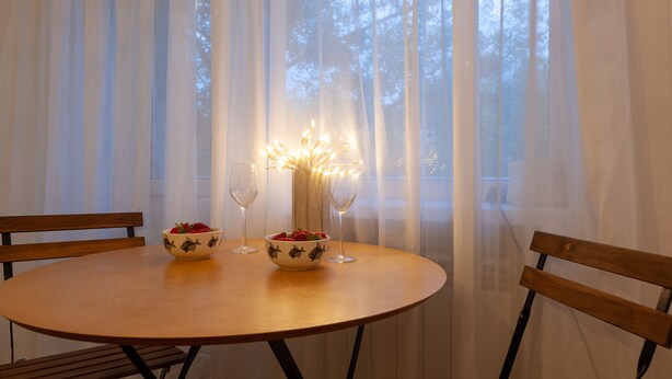 A round wooden table with two bowls of strawberries and two wine glasses, set against a backdrop of sheer curtains and a window with a view of trees. - MalinaApt  Sophisticated red (Kyiv)