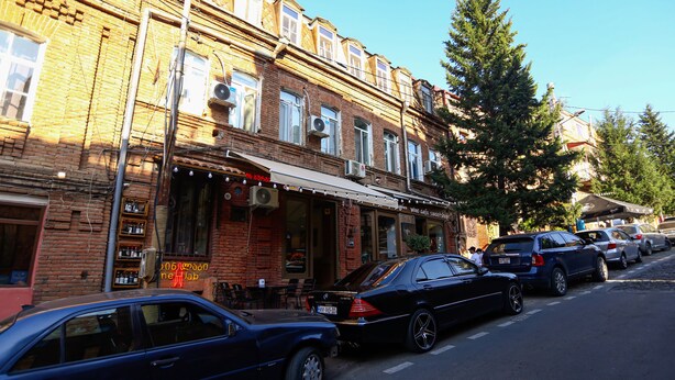 Straßenansicht mit parkenden Autos, einem Backsteingebäude mit Café und einem großen Baum.