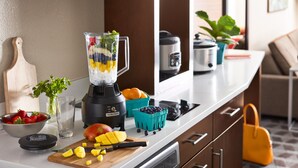 A kitchen counter with a blender, fruits, a cutting board, a bowl of strawberries, and a potted plant.