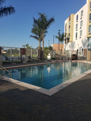 Uma piscina com elevador e sinalização de acessibilidade para cadeiras de rodas, rodeada por palmeiras e um edifício com várias janelas.