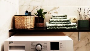 Een wasmachine met bedieningspaneel, een rieten mand, een potplant en opgevouwen handdoeken op een zwart aanrecht.