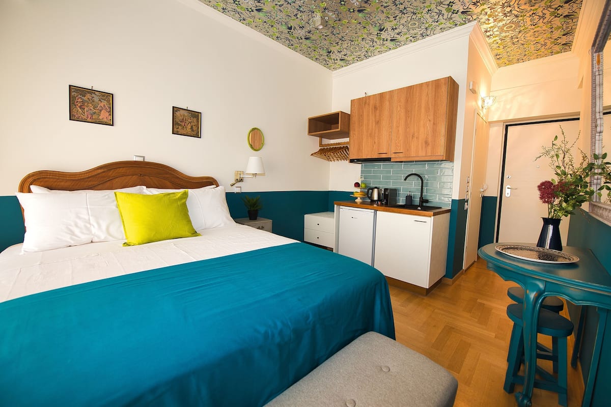 Ein Schlafzimmer mit einem Bett, einem Nachttisch, einem kleinen Tisch mit Lampe, einer Küchenzeile mit Holdekor und einer Badezimmertür.
