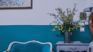 Un fauteuil recouvert d’un tissu bleu-vert, une petite table d’appoint blanche ornée d’un vase de fleurs jaunes et une lampe fixée au mur.