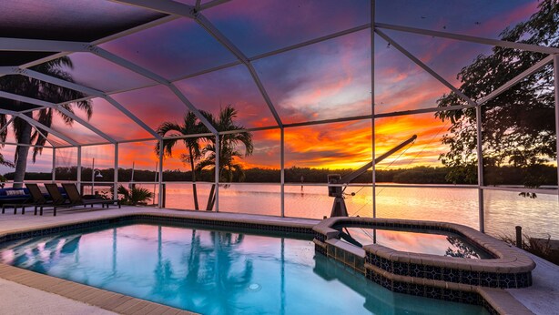 Ein Poolbereich mit Blick auf einen Sonnenuntergang über dem Wasser, umgeben von Palmen und einer überdachten Sitzgelegenheit.