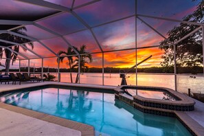 A pool area with a view of a sunset over water, surrounded by palm trees and a covered seating area. - ⚓Casa Palma! Beautiful waterfront home w/heated pool & dock!⚓🌅 (Bradenton)