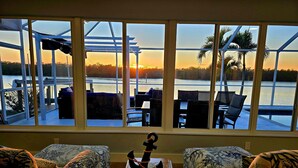 Ein Zimmer mit großem Fenster, das auf ein Gewässer und einen Sonnenuntergang blickt. Zwei gemusterte Sofas und ein kleiner Tisch mit einem dekorativen Gegenstand in der Mitte.