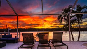 Eine Terrasse mit drei Stühlen und einer Couch, die Blick auf einen Sonnenuntergang über dem Wasser mit Palmen bietet.
