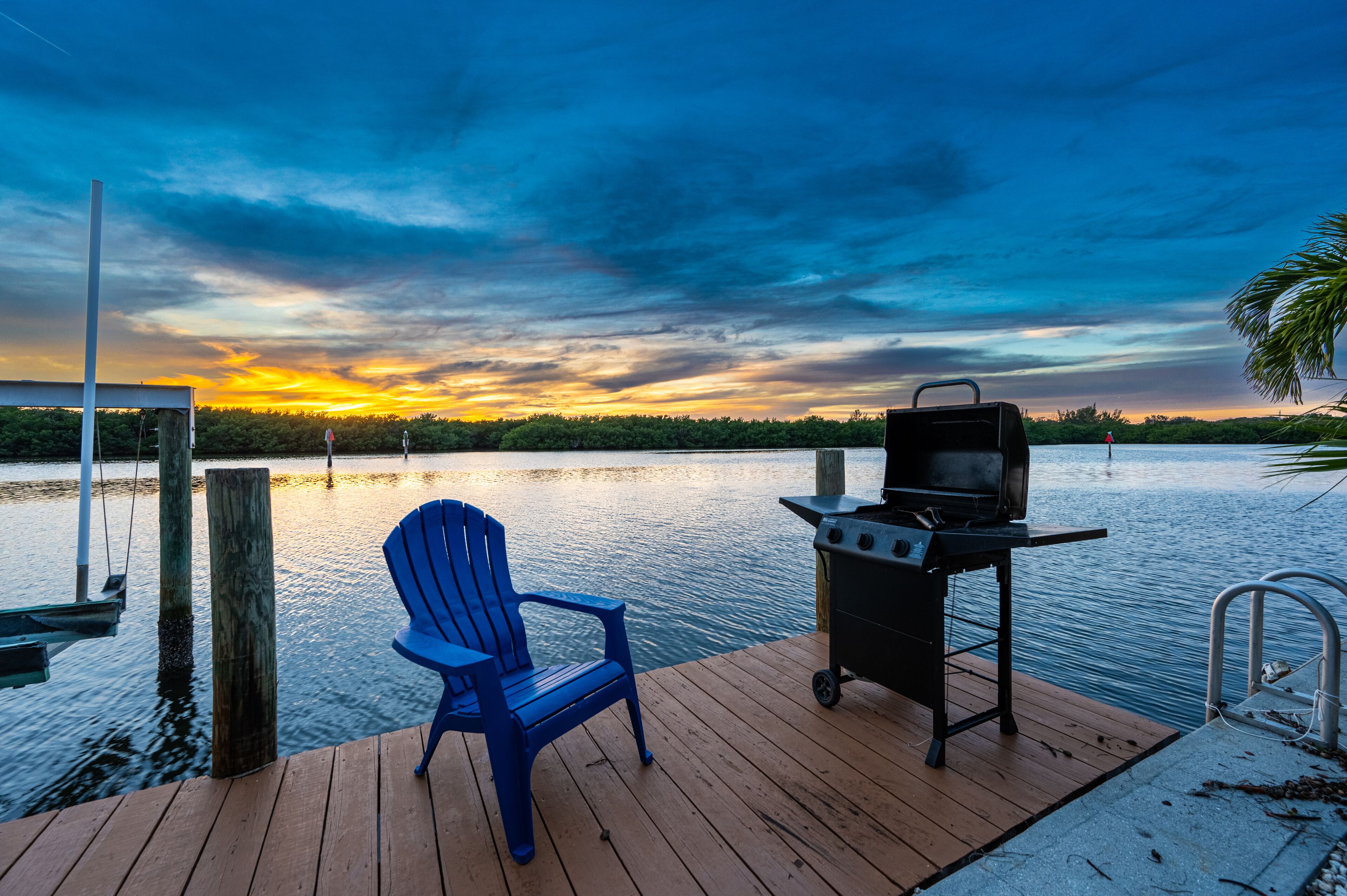 Puinen terassi, jossa on sininen Adirondack-tyylinen tuoli ja grilli. Terassilta avautuu näkymä tyyneen järveen, ja taustalla näkyy auringonlasku.