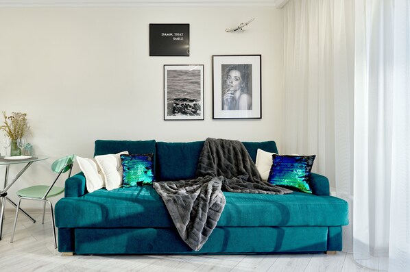 A teal sofa with a blanket, two framed pictures, and a small table with a vase and glasses.