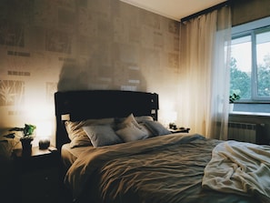 Quarto com cama, mesas de cabeceira, abajur e janela com cortinas.