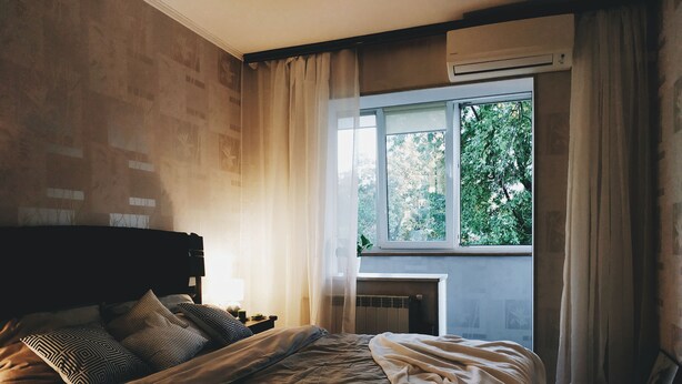 A bedroom with a bed, a nightstand, a window with a view of greenery, and an air conditioning unit. - MalinaApt Hello Geometry (Kyiv)