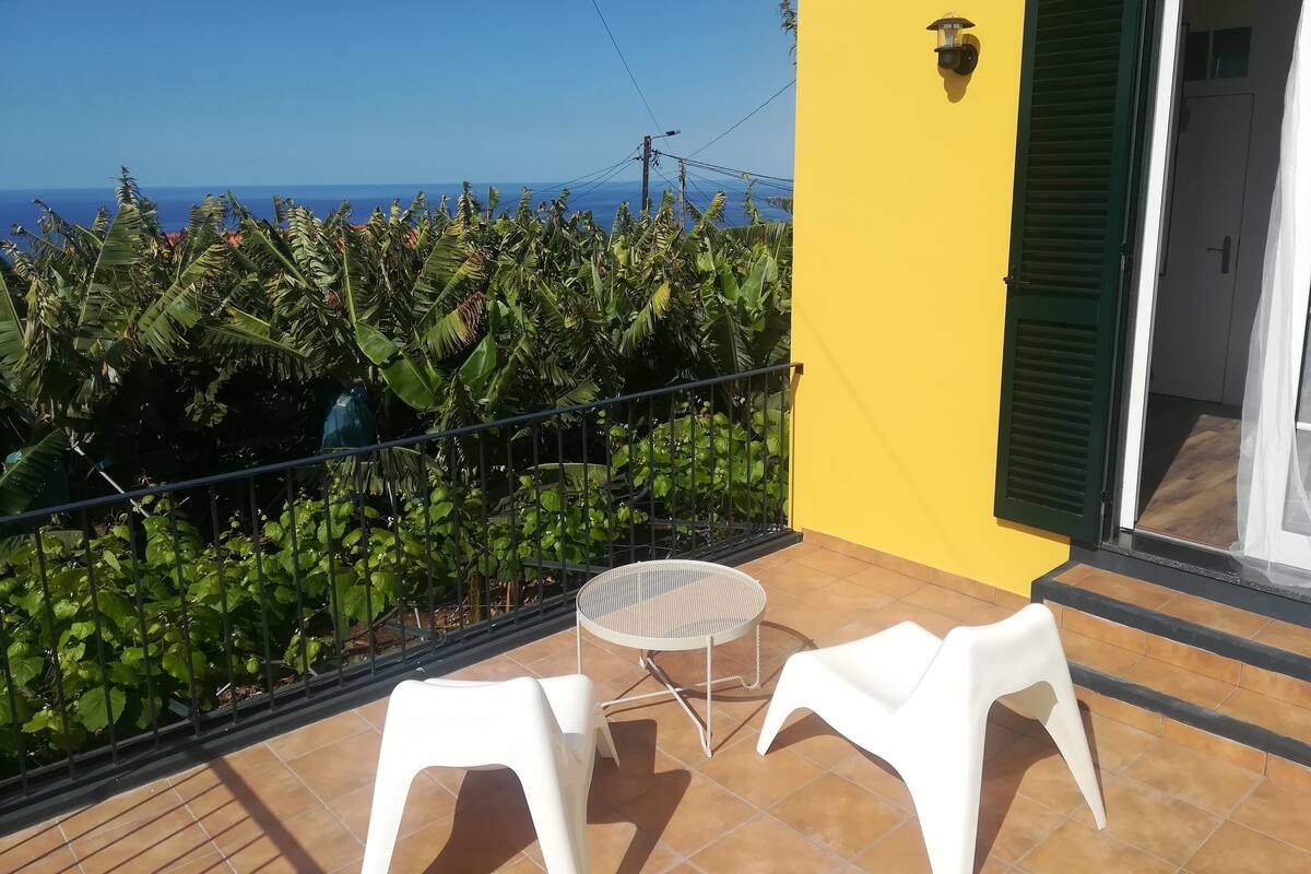 A balcony with a view of the sea, a yellow wall, and a door with green shutters.