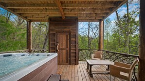Une terrasse en bois avec un jacuzzi, des fauteuils en osier et une porte en bois donnant sur une zone boisée.
