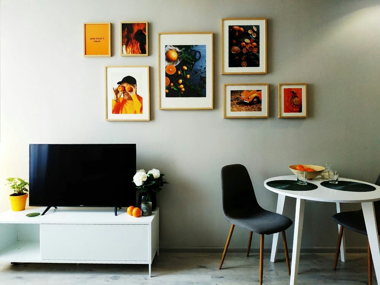 A modern living room with a flat-screen TV, a white entertainment unit, a dining table with two chairs, and a wall adorned with framed artwork.