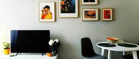 A modern living room with a flat-screen TV, a white entertainment unit, a dining table with two chairs, and a wall adorned with framed artwork.