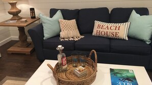A living room with a dark blue sofa, two light blue pillows, a 'BEACH HOUSE' pillow, a woven basket with a bottle and glasses, a magazine, and a lamp on a side table. - The Frankfort Surf Cottage (Frankfort)