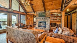 A cozy cabin interior with a stone fireplace, wooden bookshelves, and a deck with a view of the mountains. - Vista Ridge- Spectacular Views | Relaxing Hot Tub | Master on Main (Blue Ridge)