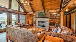 A cozy cabin interior with a stone fireplace, wooden bookshelves, and a deck with a view of the mountains. - Vista Ridge- Spectacular Views | Relaxing Hot Tub | Master on Main (Blue Ridge)