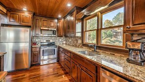 A kitchen with wooden cabinets, stainless steel appliances, granite countertops, and a large window offering a view of the outdoors. - Vista Ridge- Spectacular Views | Relaxing Hot Tub | Master on Main (Blue Ridge)