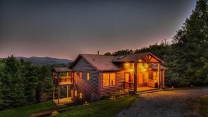 Eine Blockhütte mit Veranda und überdachter Eingangsterrasse, umgeben von Bäumen und einem Berg im Hintergrund.